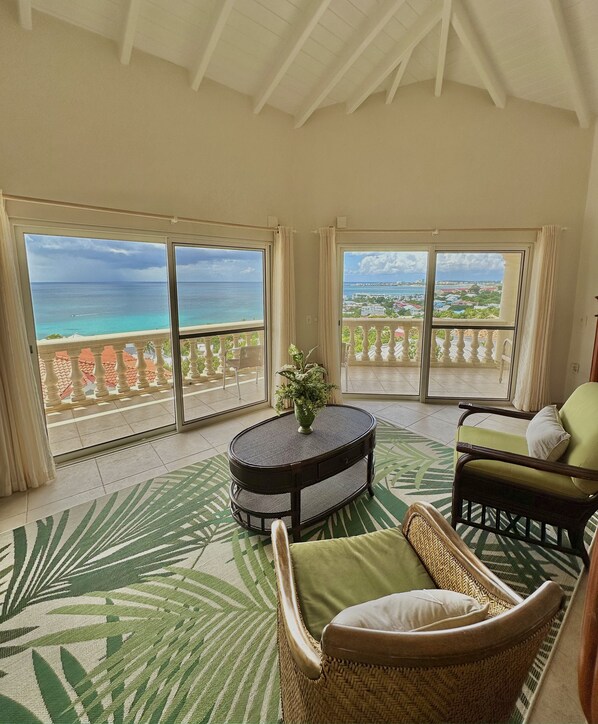 A room with a round table, a wicker chair, and a view of the ocean through large windows.