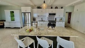 A modern kitchen with white cabinetry, a central island with seating, and a stainless steel refrigerator. - Pelican Pearl Villa - GENERATOR (Sint Maarten)