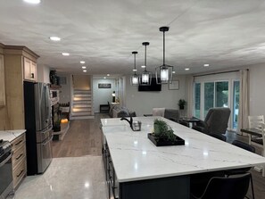 Een moderne keuken met een groot eiland, roestvrijstalen apparatuur, hanglampen en uitzicht op de buitenruimte.