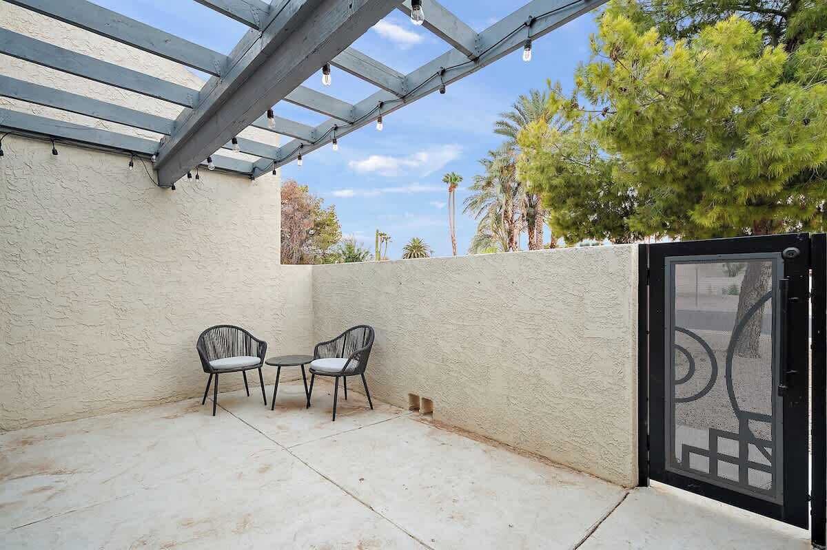 Un patio cubierto con dos sillas y una mesa, una pérgola con luces colgantes, y vistas a árboles y una reja.