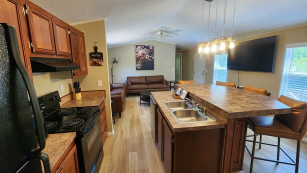 A modern kitchen with wooden cabinets, a black refrigerator, a built-in oven, a granite countertop, a double sink, and a dining area with a brown leather couch and a table. - Bunk house on the ranch (Tyler)