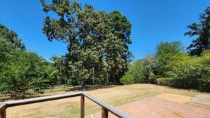 A wooden deck with a metal railing, a paved area with a patterned surface, and a grassy field with trees in the background. - Bunk house on the ranch (Tyler)