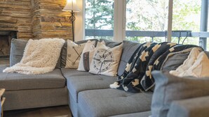 A cozy living room with a stone fireplace, a sofa with patterned cushions, and a view of trees through large windows.