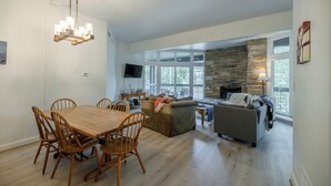 A spacious living area with a dining table, wooden chairs, a stone fireplace, and a large window.