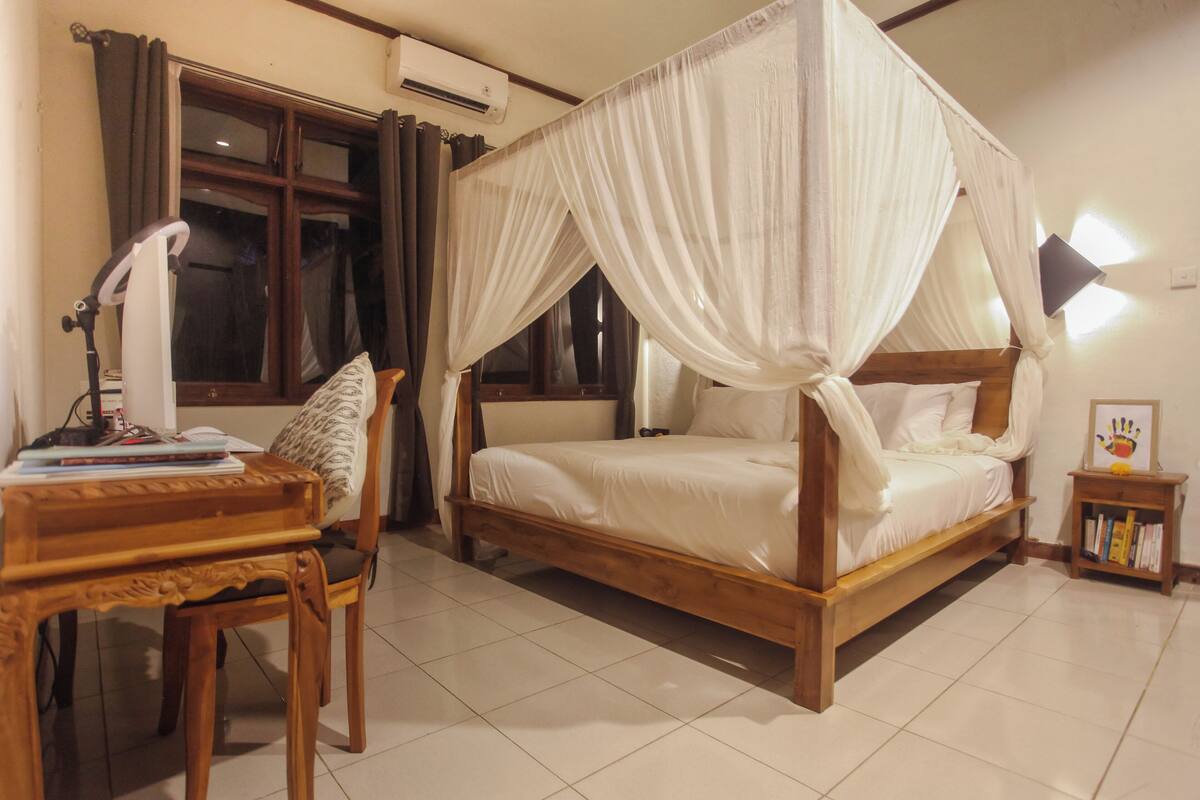 A bedroom with a four-poster bed, a wooden desk with a chair, a bedside table with a lamp, and a small bookshelf.
