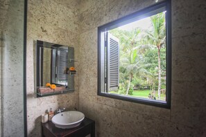 Een badkamer met een marmeren wand, een groot raam met uitzicht op groen, een spiegel en een wastafel met toiletartikelen.