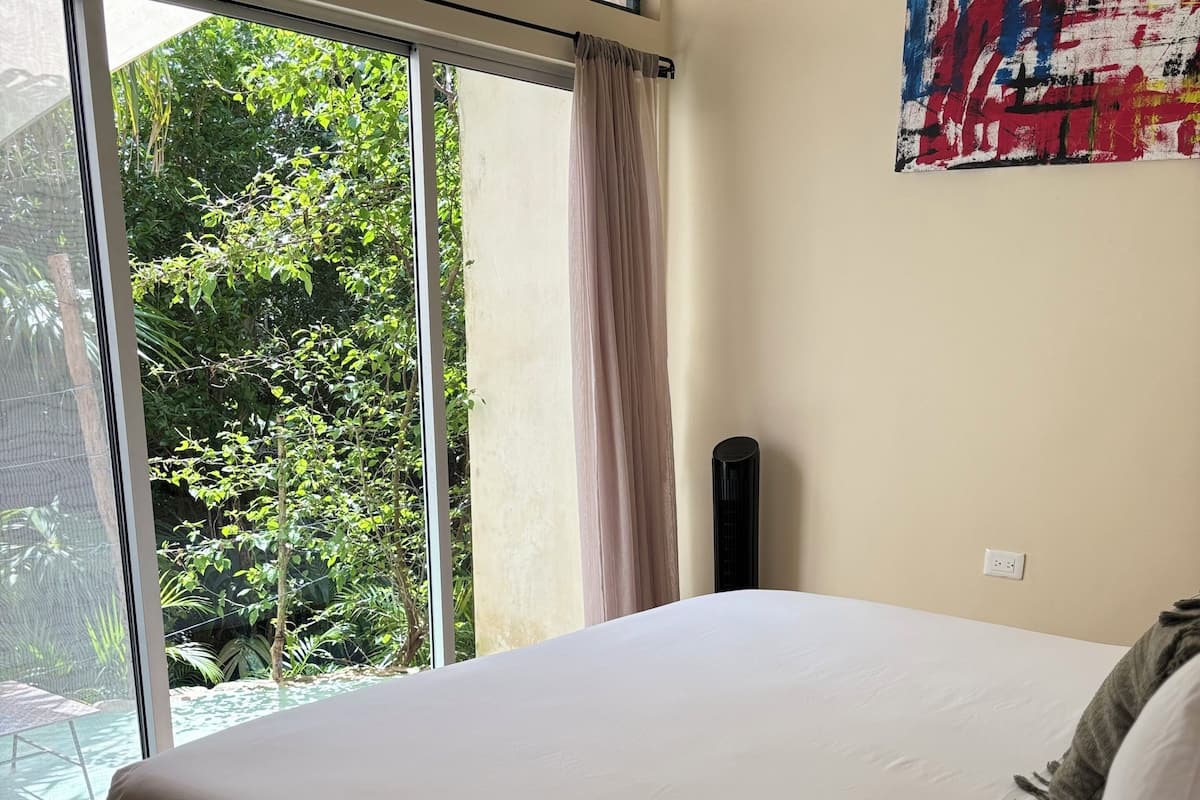 Ein Schlafzimmer mit einem großen Bett, Blick ins Grüne durch eine Schiebetür, einem Bild an der Wand und einem Nachttisch mit Lampe.