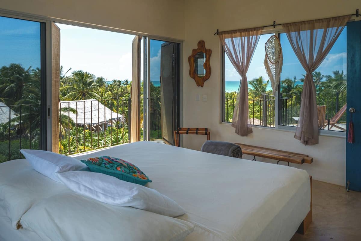 Ein Schlafzimmer mit einem großen Bett, einer Bank, einem Sessel und Blick auf Palmen und das Meer durch offene Fenster.