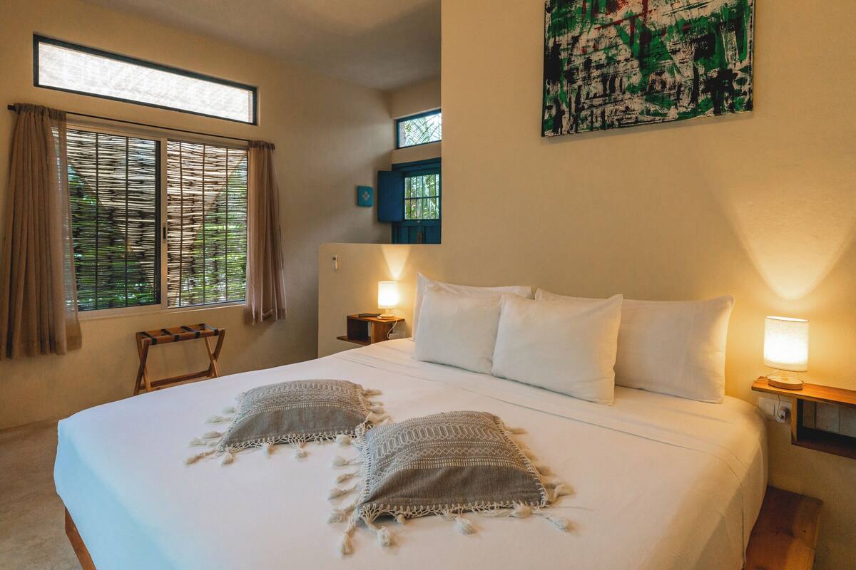 Ein Schlafzimmer mit einem großen Bett, zwei Nachttischlampen, einem Fenster mit Vorhängen, einem kleinen Tisch und einem Bild an der Wand.