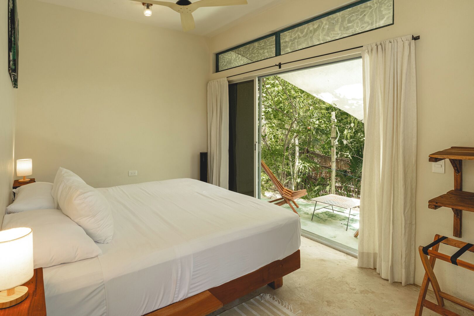 A bedroom with a bed, a nightstand, a lamp, a ceiling fan, a window with curtains, and a view of greenery outside.