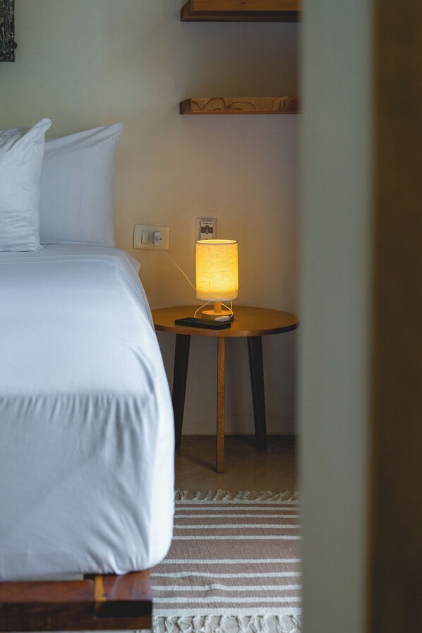 A neatly made bed with white linens, a small round table with a lamp, and a power outlet on the wall.
