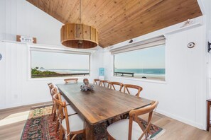 A dining room with a wooden table and chairs, large windows with ocean views, and a hanging light fixture.