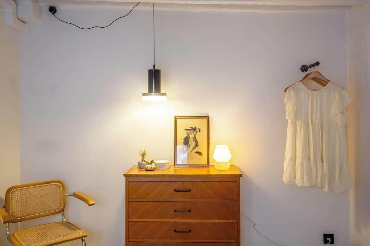 Ein Zimmer mit einem hölzernen Schminktisch, einem Korbstuhl, einer Pendelleuchte, einem gerahmten Bild und einem weißen Kleid an einem Kleiderbügel.