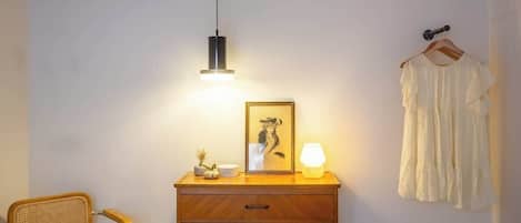 A room with a wooden dresser, a wicker chair, a hanging light, a framed picture, and a white dress on a hanger.