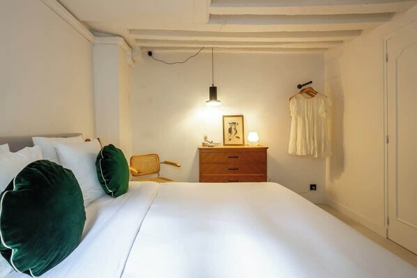 Un lit bien fait, avec du linge de lit blanc et des coussins verts, une table de chevet en bois avec une lampe, un tableau encadré et un vêtement blanc accroché au mur.