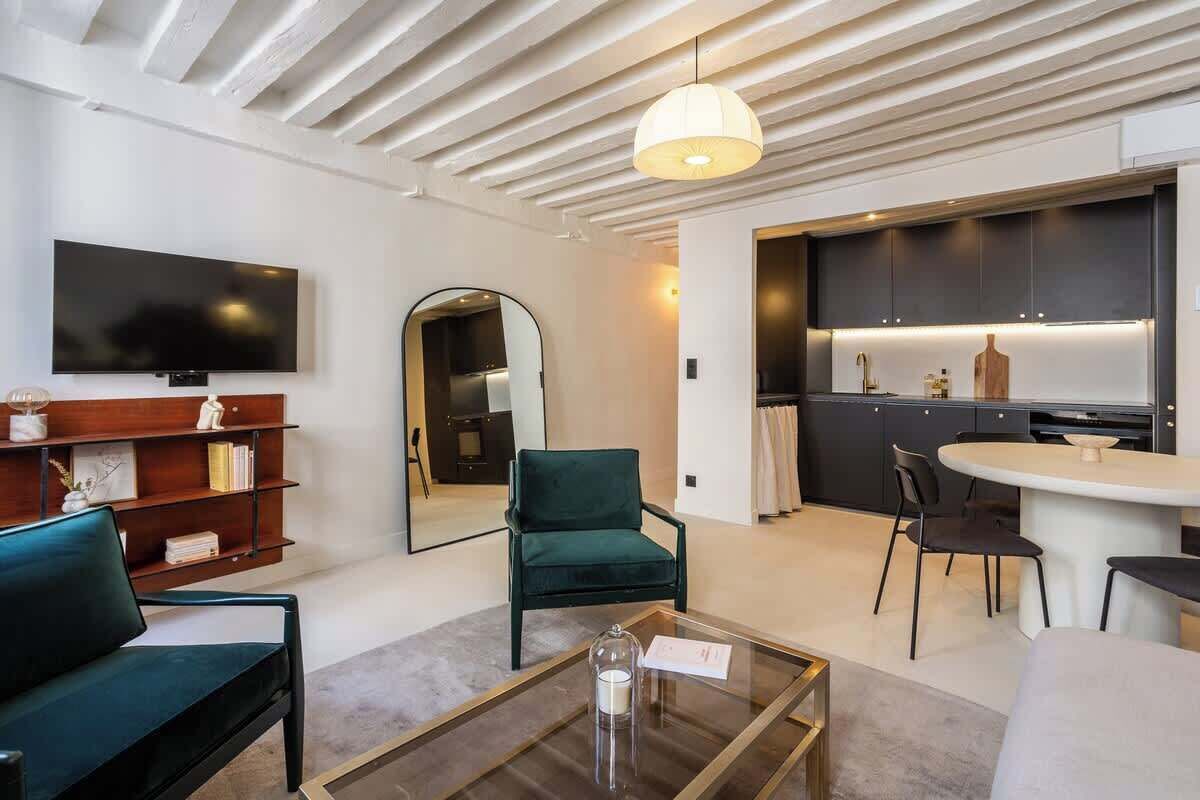 A modern living room with a sofa, a coffee table, a TV, a bookshelf, a green armchair, and a kitchen area with dark cabinets and a round dining table.