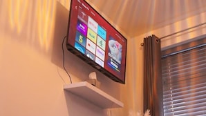 Ein Schlafzimmer mit einem Bett, einem an der Wand montierten Fernseher auf einem Regal darüber und einem Fenster mit Jalousien.