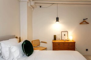 Una cama bien tendida con sábanas blancas, un aparador de madera con cajones, una silla de mimbre y una lámpara colgante.