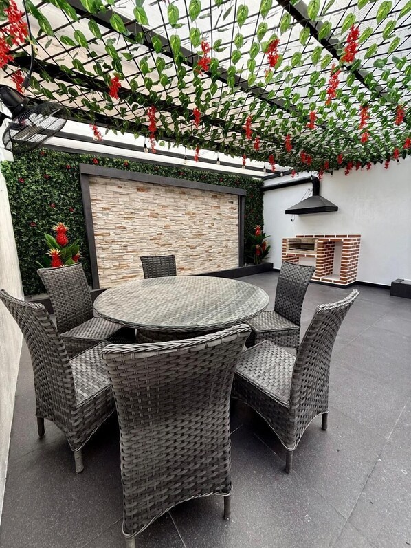 A round outdoor dining area with wicker chairs and a table, surrounded by a brick wall and greenery.