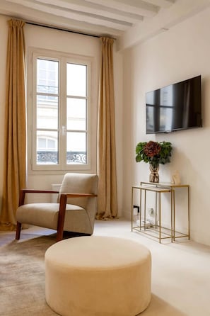 Uma sala de estar moderna com uma poltrona bege, um banquinho redondo, uma mesinha lateral com um vaso de flores e uma televisão de tela plana na parede.