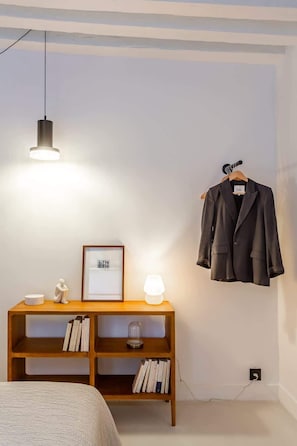 Quarto com cama, mesa de cabeceira de madeira com livros, quadro emoldurado, casaco pendurado e luminária pendente.