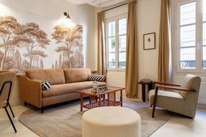 Uma sala de estar com sofá, poltrona, mesa de centro e um banquinho. Há uma janela grande com cortinas, um mural na parede e um quadro emoldurado.