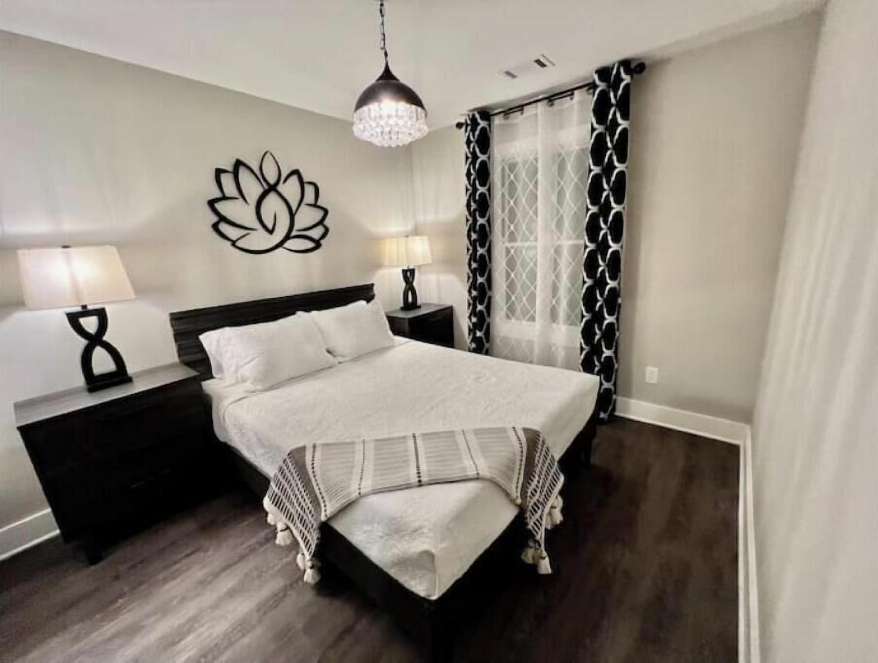 Une chambre à coucher avec un grand lit, deux tables de chevet avec des lampes, un élément décoratif mural et une fenêtre agrémentée de rideaux à motifs.