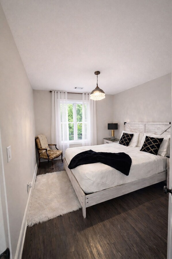 A bedroom with a white bed, dark wooden flooring, a white chair, and a window with curtains.