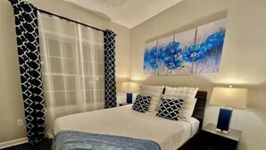 A bedroom with a bed, two bedside tables, a large window with curtains, and a painting on the wall.