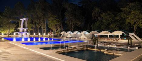 A nighttime swimming pool area with a waterfall feature, seating areas, and a covered pavilion.