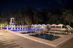 Uma área de piscina à noite, com uma cascata, espaços para sentar e um pavilhão coberto.