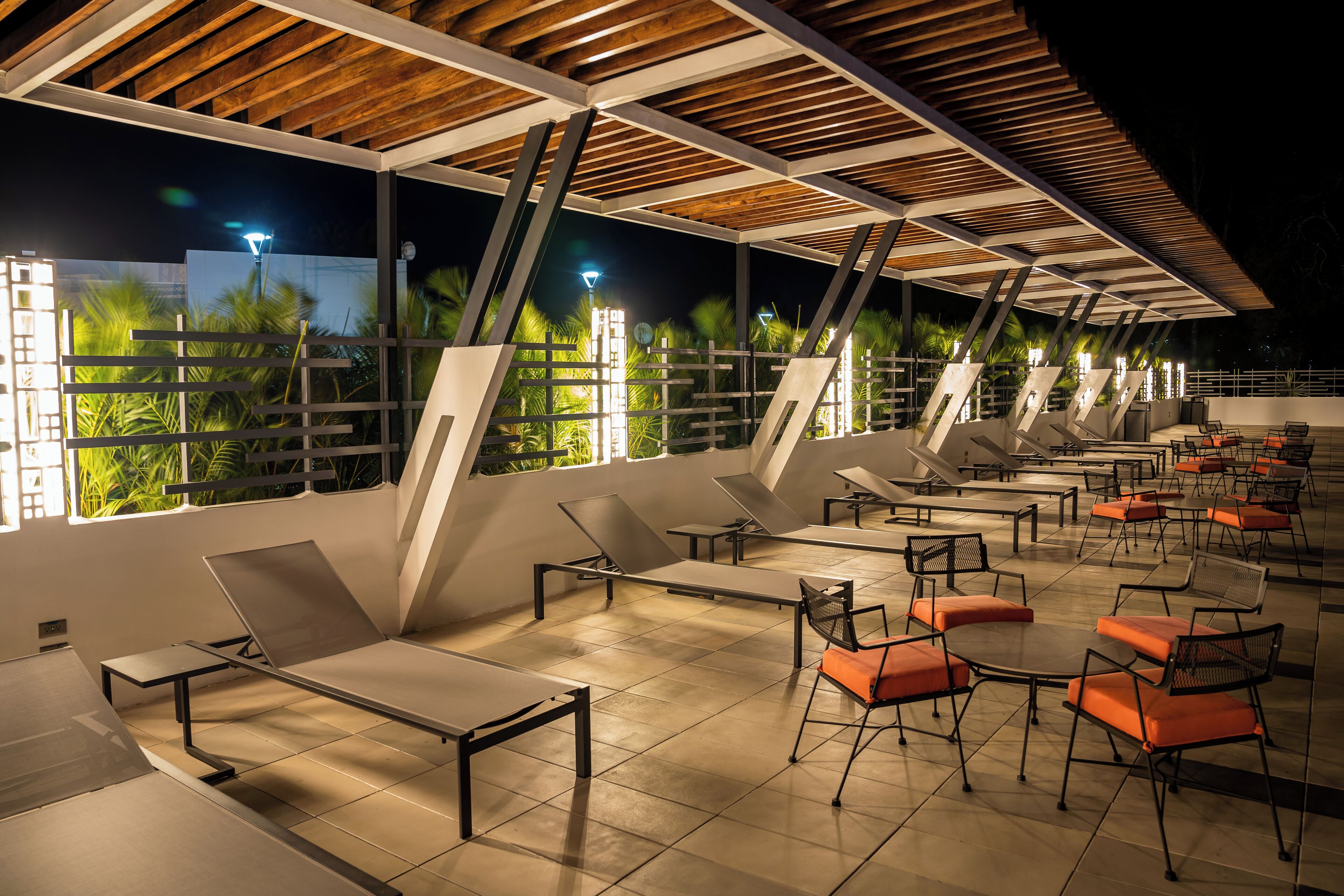 An outdoor lounge area with sun loungers and tables, large windows, and a wooden ceiling structure.