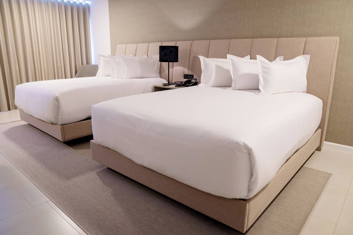A hotel room with two beds, a bedside table with a lamp, and a view of the outside through curtains.