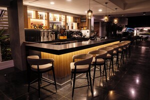 Uma área de bar moderna com um balcão comprido, vários bancos e uma variedade de garrafas e copos em prateleiras.