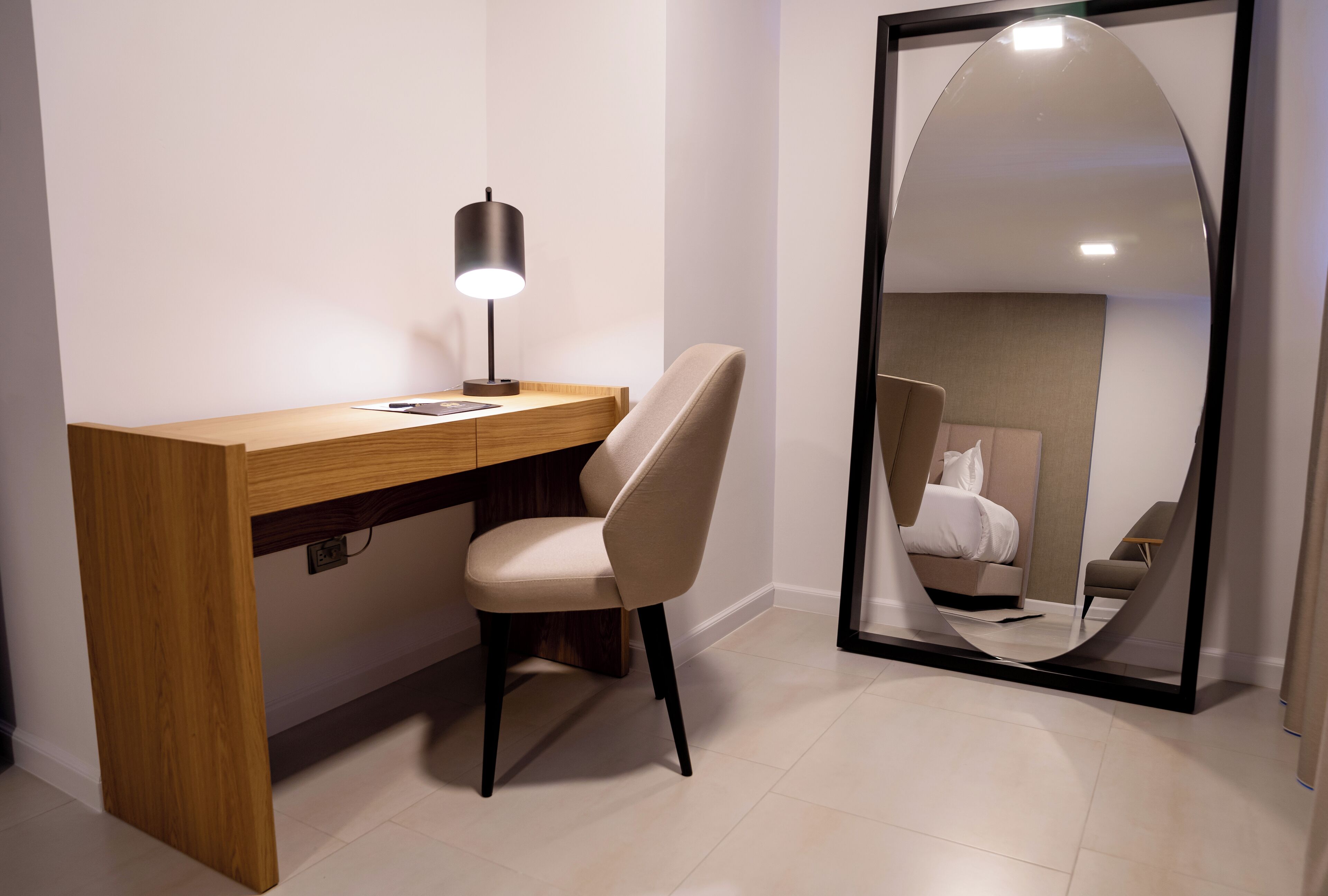 Habitación de hotel moderna con escritorio de madera, una silla, lámpara de escritorio y un espejo grande que refleja la cama y una silla.