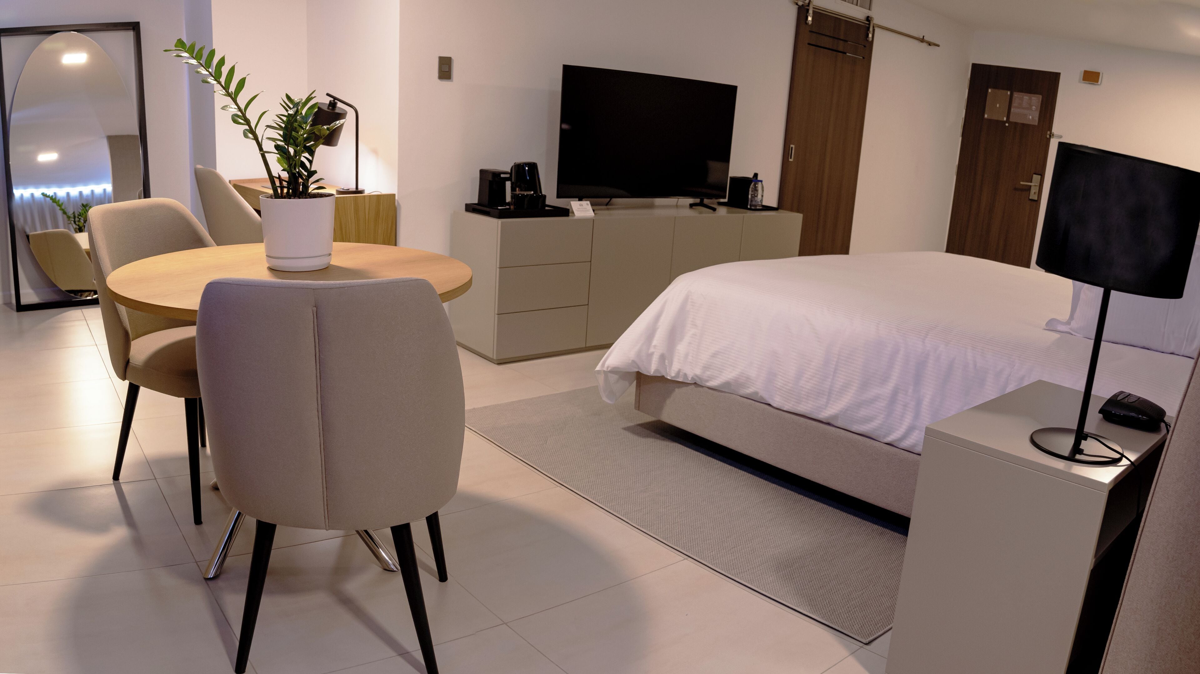 A modern hotel room with a bed, a desk, a chair, a television, a coffee maker, and a plant.
