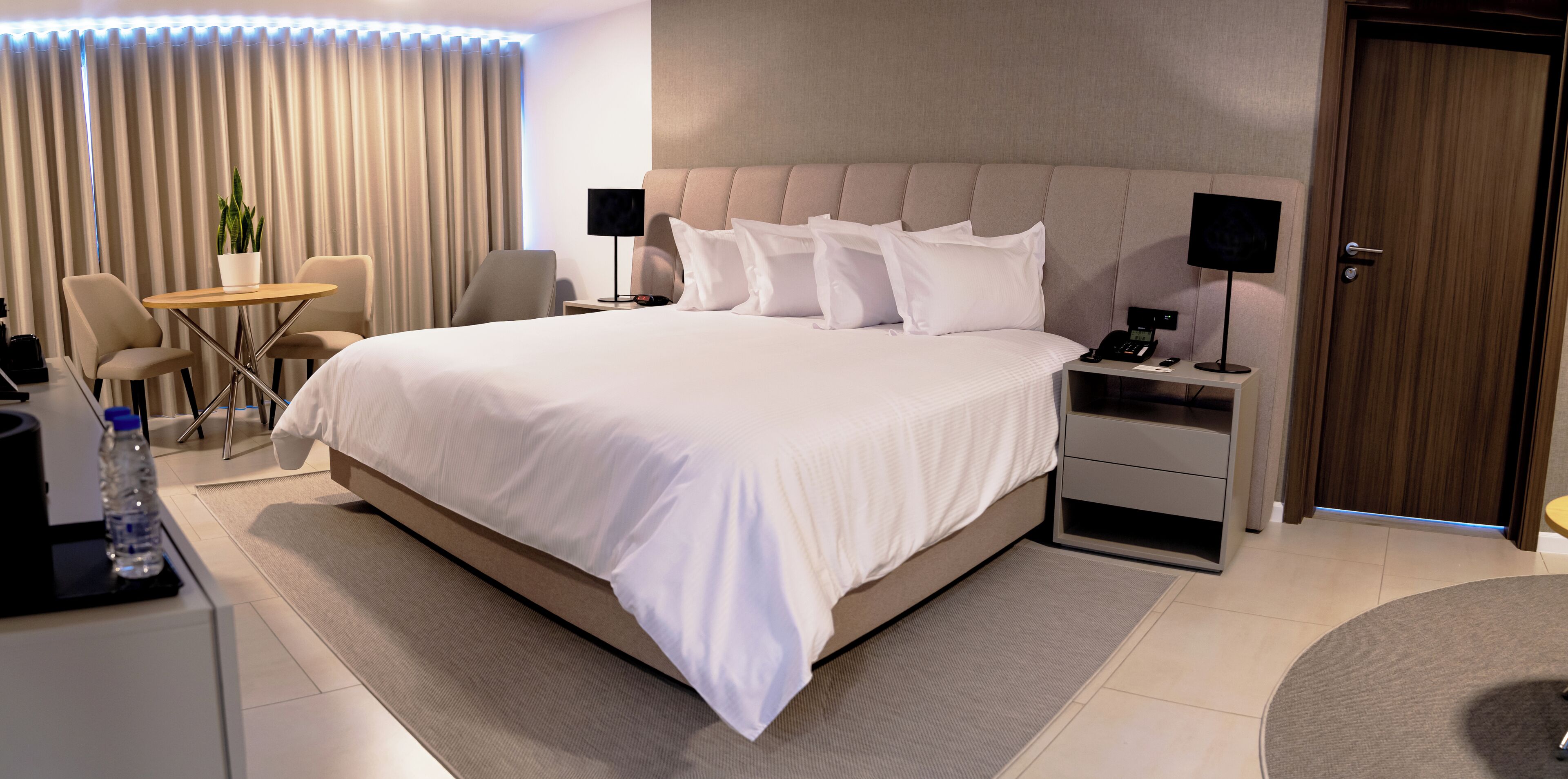 A modern hotel room with a large bed, bedside tables, a desk with a chair, and a potted plant.