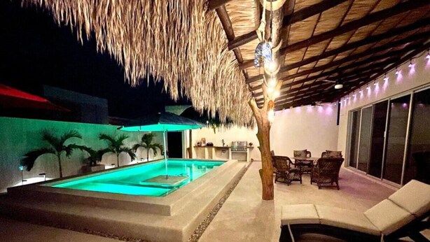Un área de piscina con techo de paja, sillones de descanso y zona de comedor.