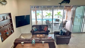 Una sala de estar con dos sofás de cuero, una mesa de centro y un televisor de pantalla plana. Hay una puerta de cristal que da a una zona de piscina con mobiliario exterior.