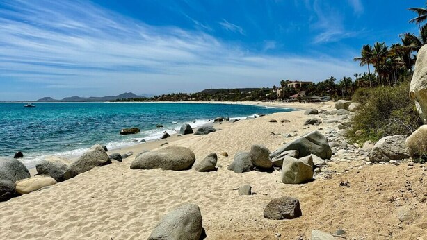 Una playa con rocas grandes, agua azul cristalina y una vista distante de edificios y montañas.