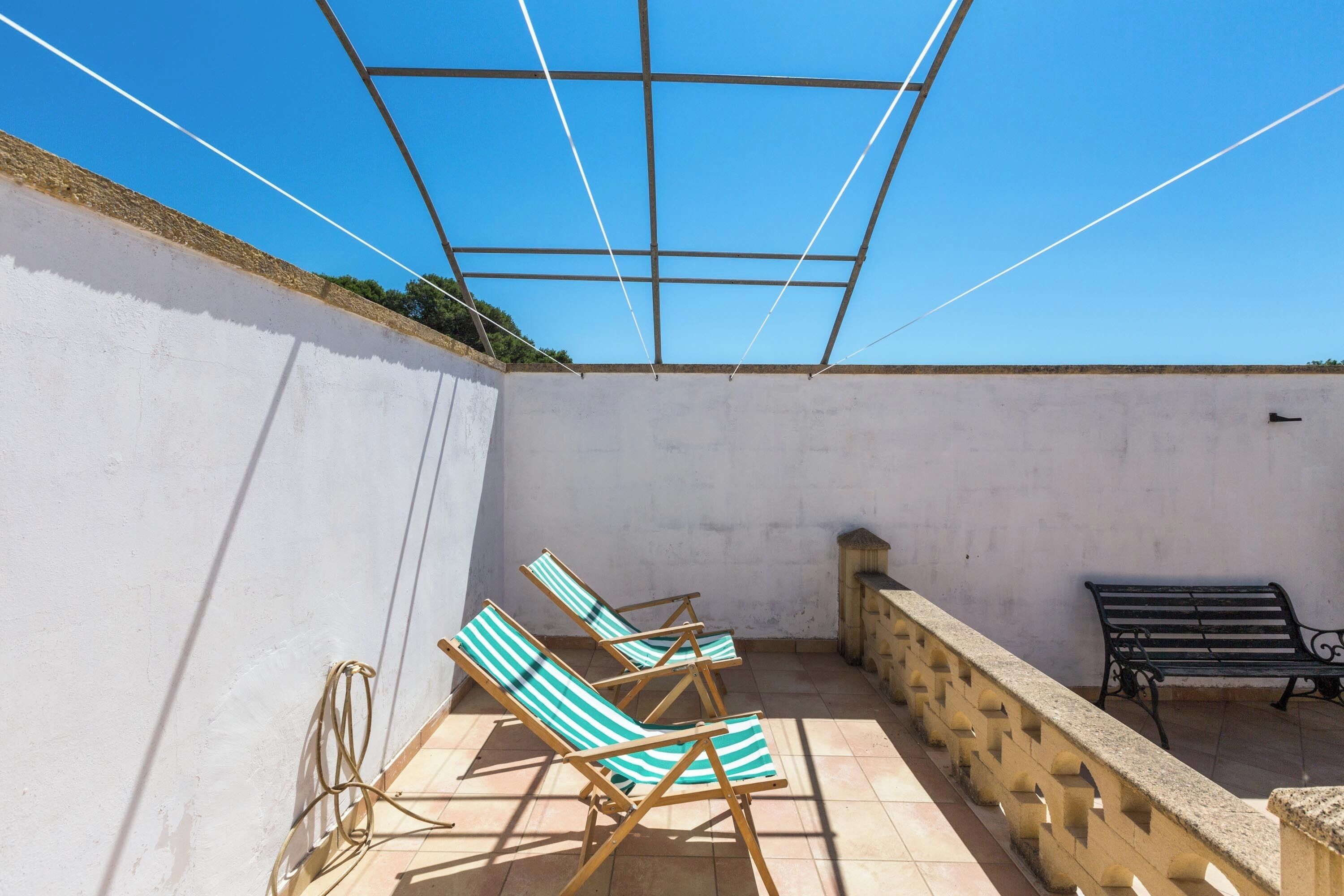 A sunlit patio with three striped lounge chairs, a stone bench, and a metal railing.