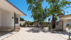 A courtyard with tiled flooring, a white building with a window, and a tree providing shade. - 4173 Villetta Ambra by Barbarhouse (Porto Cesareo)