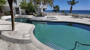 A pool with a curved design, surrounded by a white building, palm trees, and a view of the ocean.