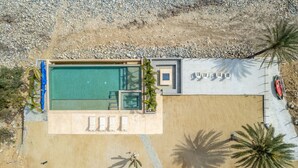 An aerial view of a modern pool area with a rectangular pool, a small adjacent structure, and a sandy area with lounge chairs and palm trees.