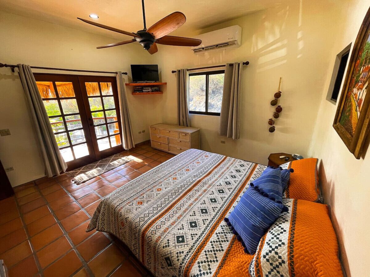 Une chambre à coucher comprenant un lit, un ventilateur de plafond, un appareil de climatisation, une télévision et une fenêtre avec des rideaux.