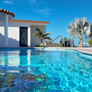 Uma piscina com água azul cristalina, uma palmeira e um edifício com parede estampada e uma porta.