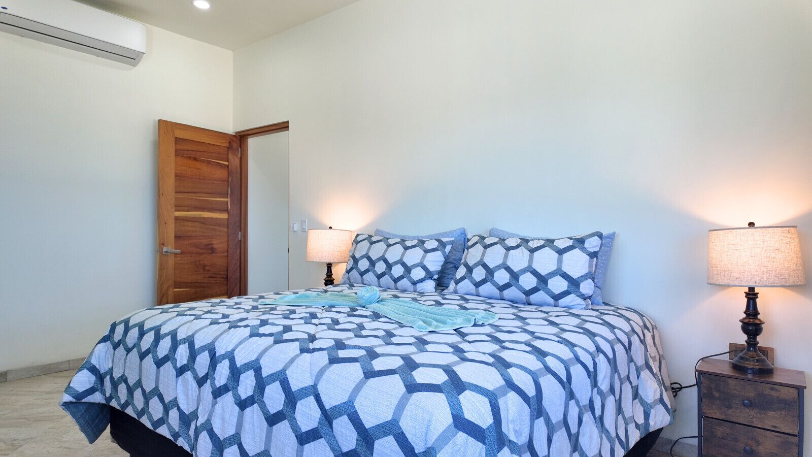 A bedroom with a bed, two bedside lamps, a wooden door, and a wall-mounted air conditioner.