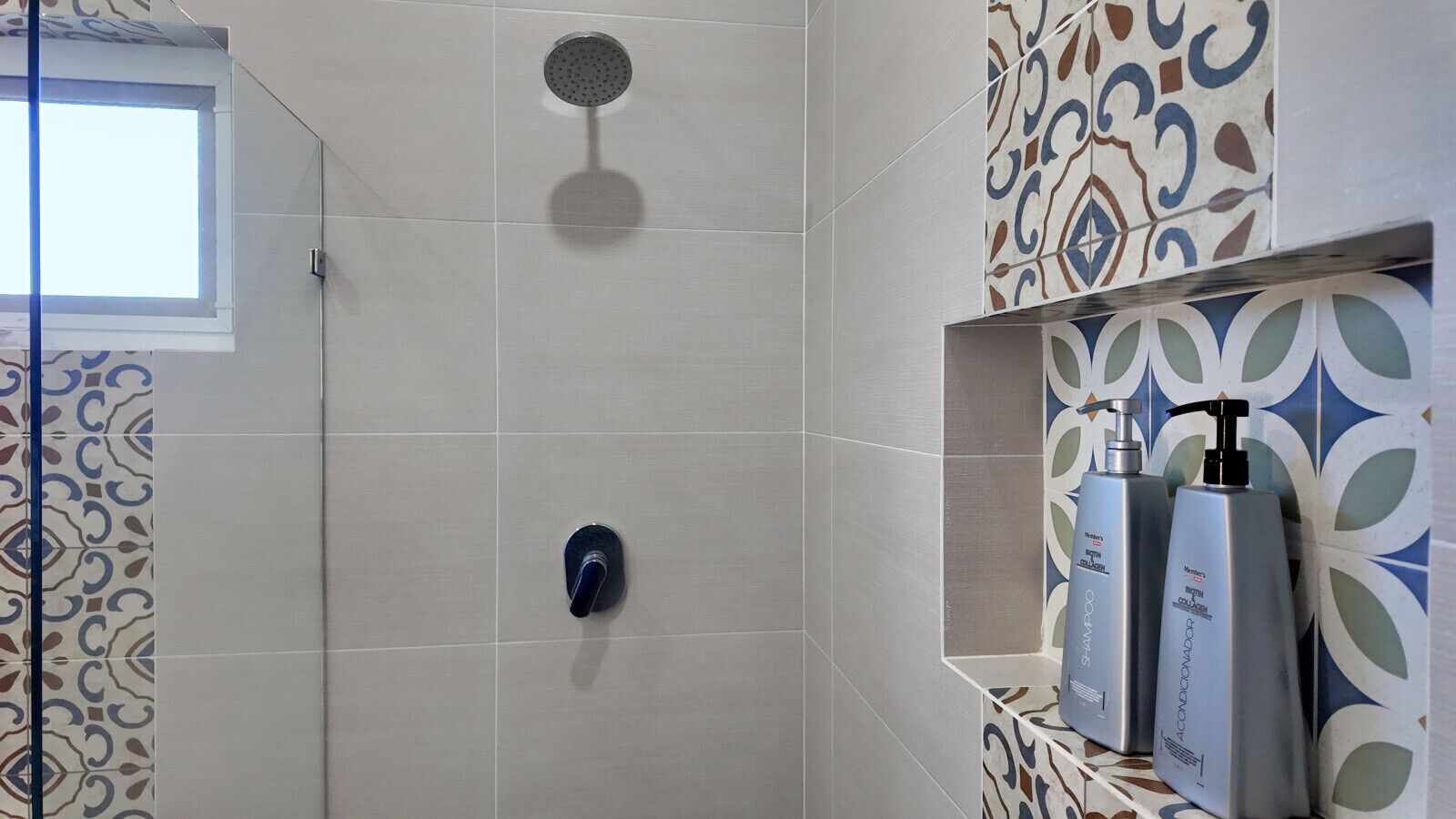 Doccia con soffione a mano e rubinetto montato sulla parete. Su uno scaffale, due flaconi di shampoo e balsamo.