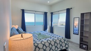 A bedroom with a bed, a lamp, a window with curtains, and a view of the sea.