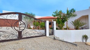 Uma entrada protegida por portão com detalhes em metal decorativo, um edifício branco com telhado de telhas vermelhas e um caminho de acesso de cascalho.